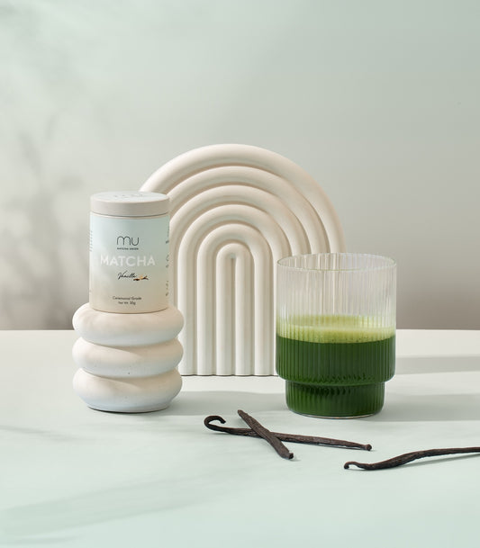 Matcha Union Vanilla Matcha tin on a white pedestal beside a ribbed glass of green matcha and vanilla pods, with a white arch backdrop.
