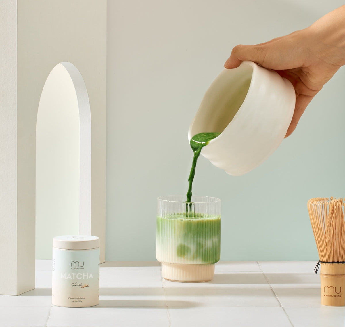 Hand pouring green matcha from a white ceramic bowl into a glass of iced milk, making a vanilla matcha latte beside a Matcha Union tin and bamboo whisk.