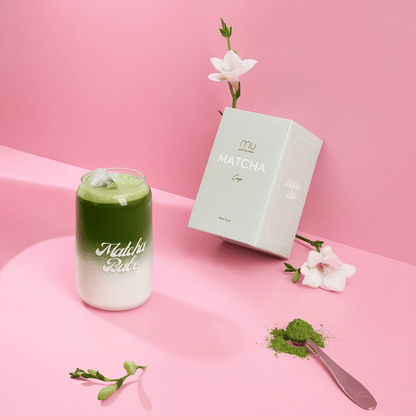 Matcha Babe glass filled with a layered iced matcha latte, beside its box, white flowers and a spoon of matcha powder on a pink display
