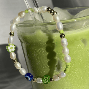 Close-up of the Matcha Magic bracelet draped over an iced matcha glass, showing freshwater pearls, gold beads, green flowers and a blue eye bead.
