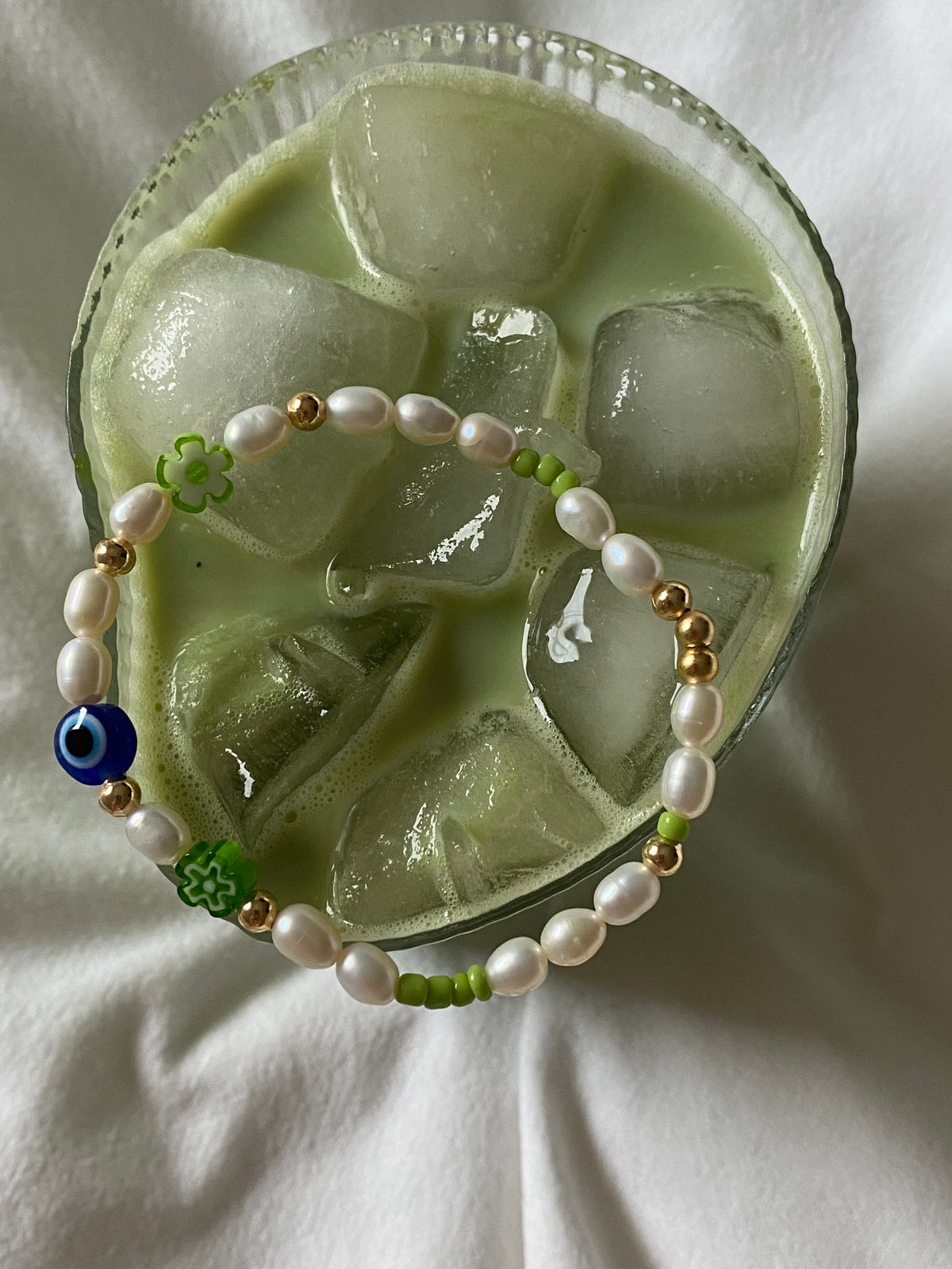 Overhead view of the Matcha Magic bracelet resting across an iced matcha glass, showing its freshwater pearls, gold beads and colourful glass details.