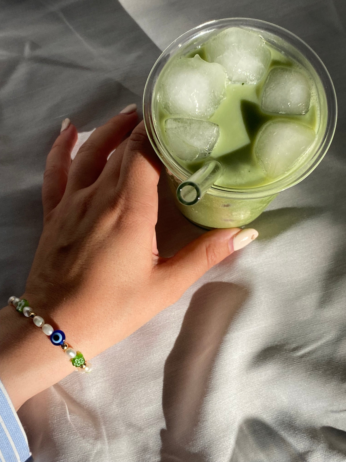 Wrist wearing the handcrafted Matcha Union x Oh Luna pearl and glass bead bracelet while holding a tall iced matcha latte with a glass straw.