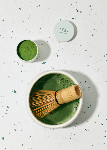 Overhead view of frothy matcha in a white ceramic bowl with a bamboo whisk, beside an open Matcha Union tin of green tea powder and its lid.