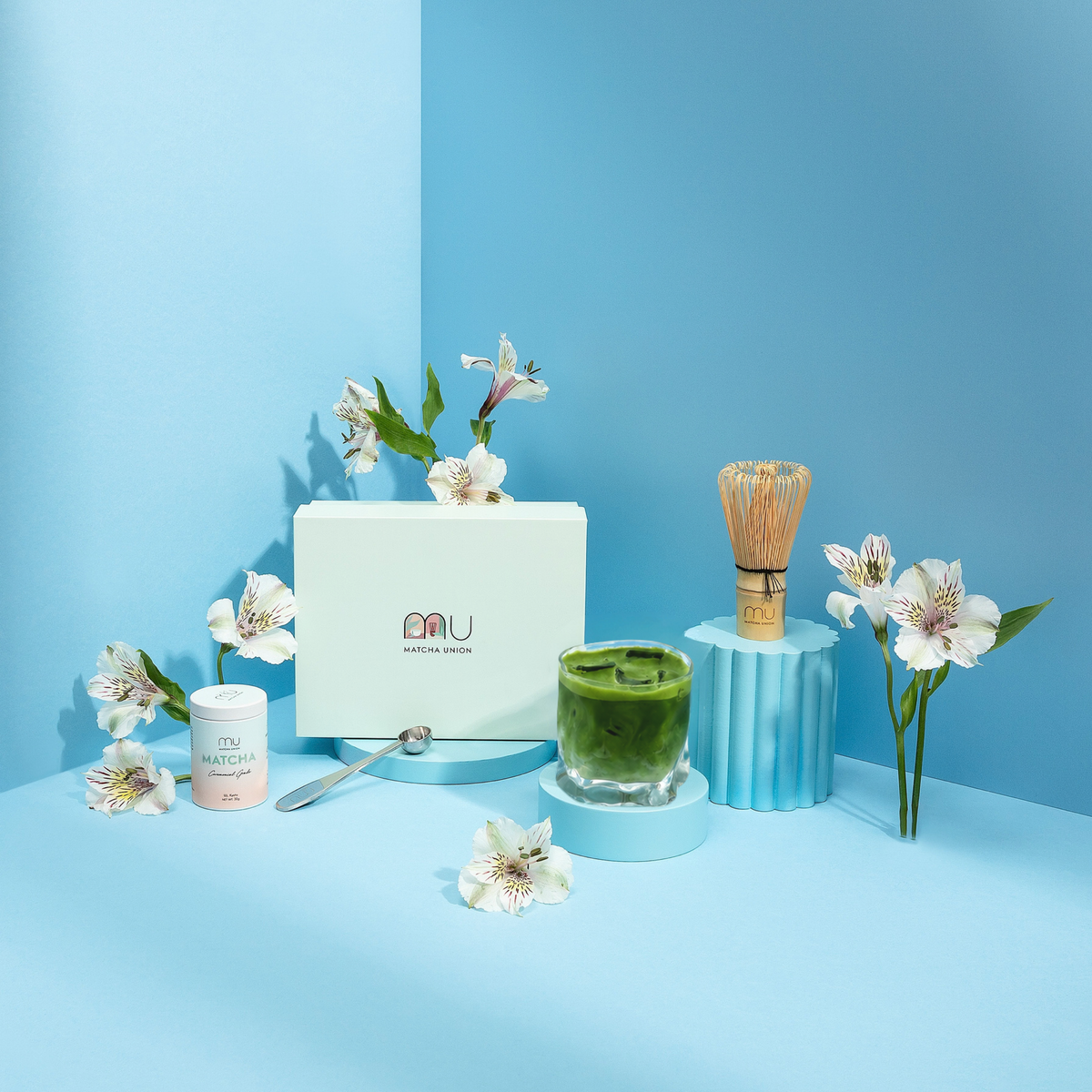 Matcha Union Ritual Set box beside a ceremonial matcha tin, stainless steel scoop, bamboo whisk and glass of iced matcha on blue stands with white flowers.
