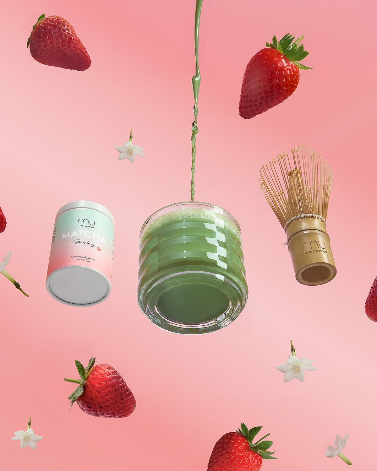 Green matcha pouring into a ribbed glass beside a Matcha Union Strawberry Matcha tin and bamboo whisk, surrounded by strawberries on a pink background.