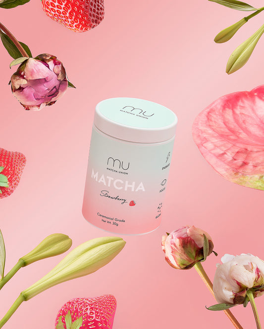 Matcha Union Strawberry Matcha tin surrounded by strawberries, pink flowers and green buds against a pink background.