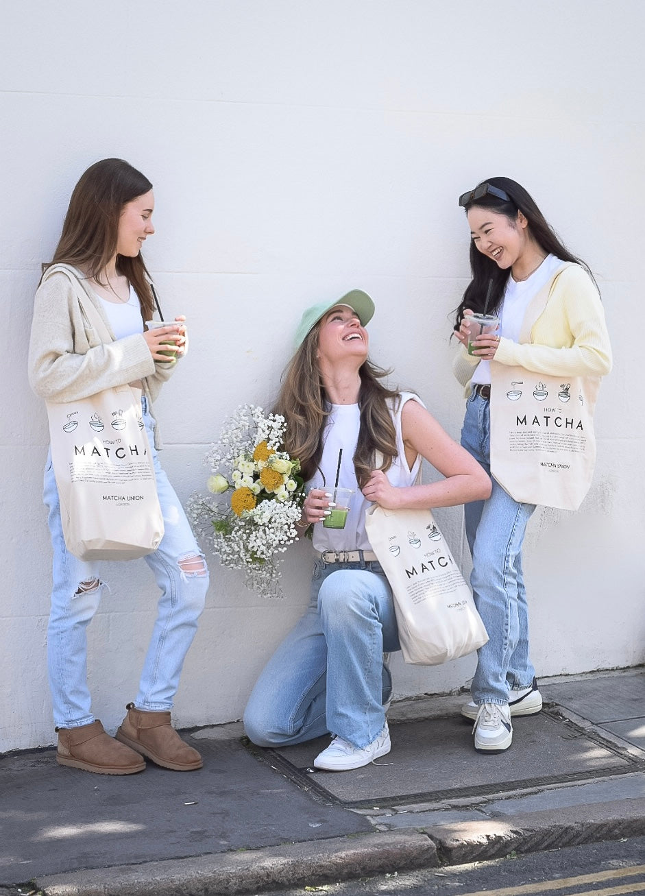 Three people carrying cream Matcha Union tote bags with illustrated matcha-making instructions while holding drinks outside against a white wall.