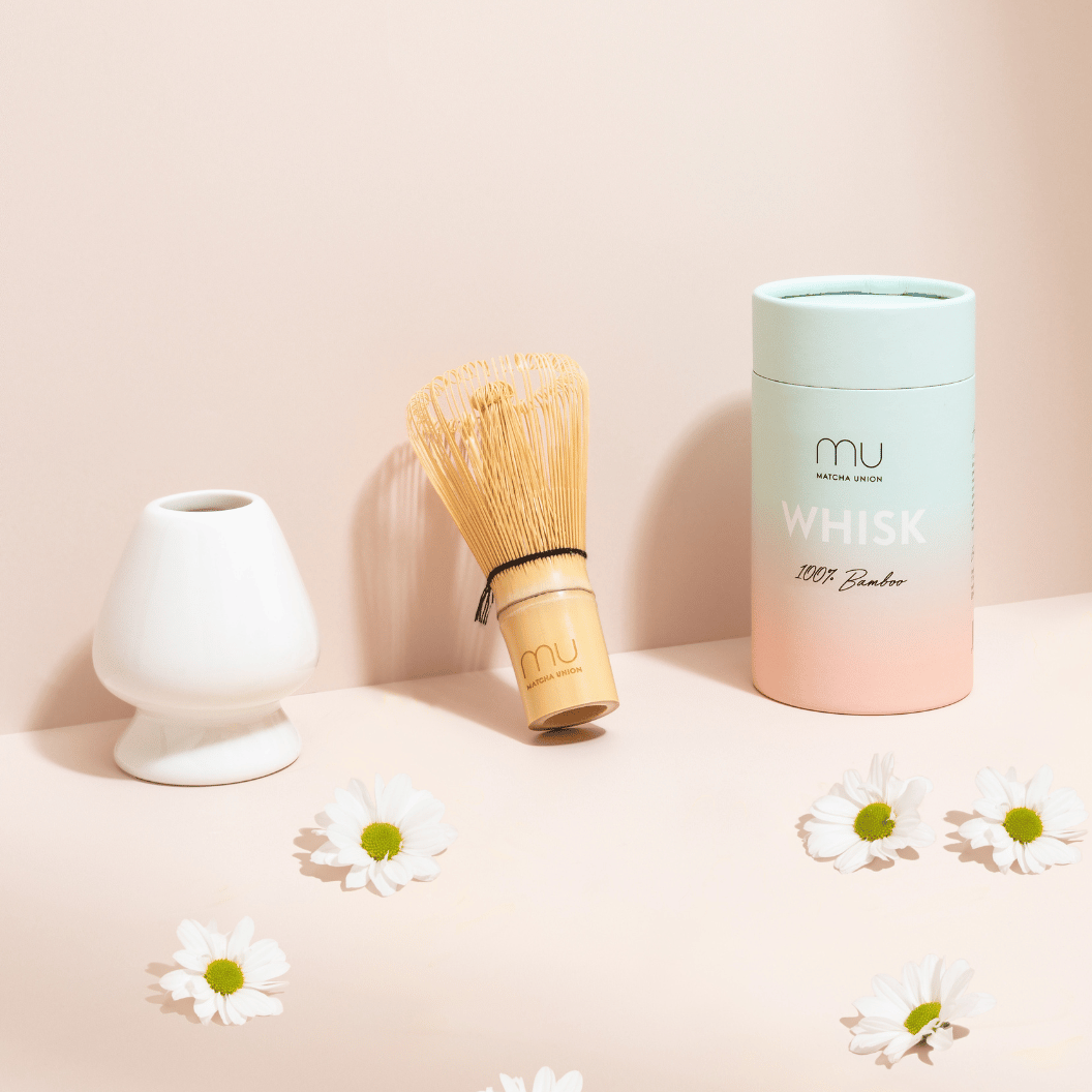 Matcha Union bamboo whisk beside a white ceramic holder and pastel whisk tube, with white daisies on a peach-coloured display.