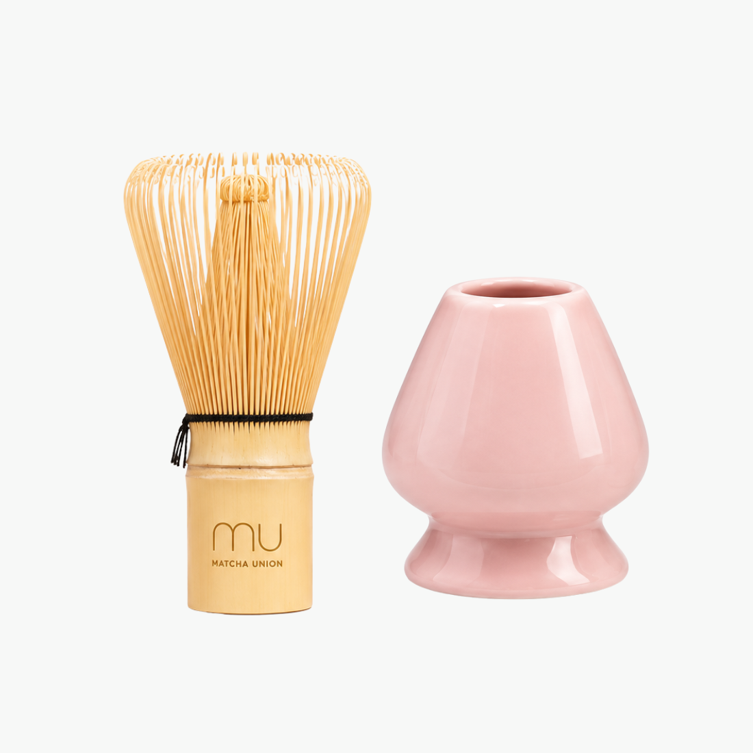 Matcha Union logo-engraved bamboo whisk beside a pink glazed ceramic holder, shaped to support the whisk’s prongs while drying.