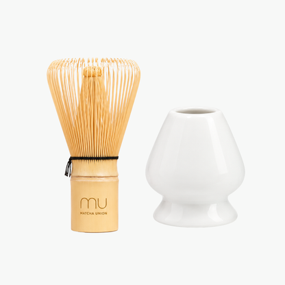 Matcha Union logo-engraved bamboo whisk beside a white glazed ceramic holder, shaped to keep the whisk’s prongs in form between uses.