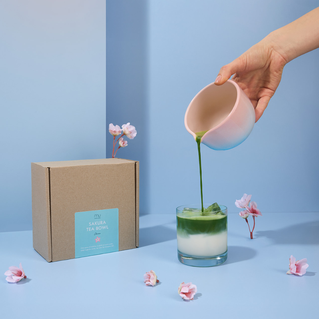 Hand pouring green matcha from the Sakura stoneware bowl into a glass of iced milk, beside its box and pink blossoms on a blue display.