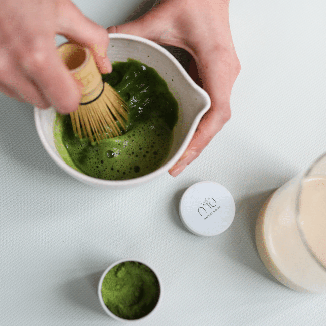 Overhead view of hands using a bamboo matcha whisk to blend green matcha into a frothy drink in a white spouted bowl.